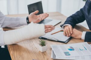 cropped view of worker with broken arm siting at table with documents opposite businessman in