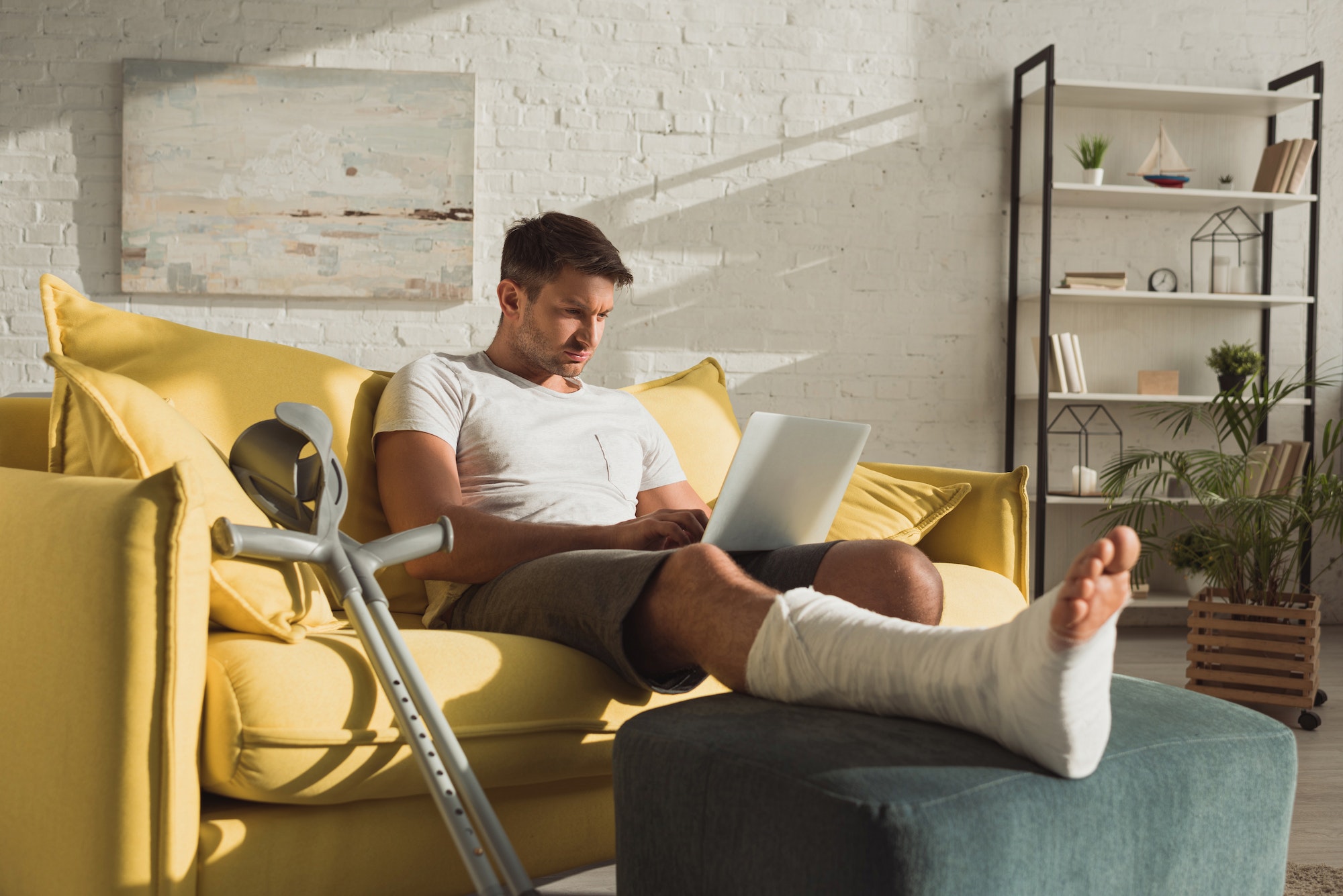 Handsome man with broken leg using laptop near crutches on sofa